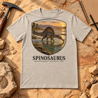 Spinosaurus t-shirt flat lay – heather dust PaleoThreads paleo art tee on sandstone with a rock hammer