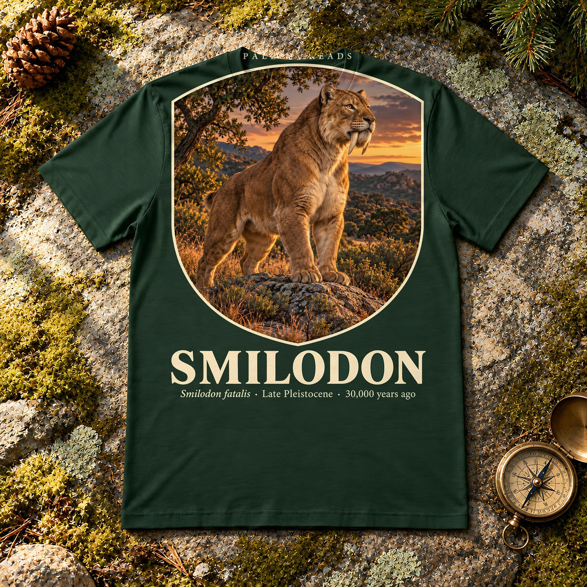 Smilodon t-shirt flat lay – forest PaleoThreads paleo art tee on mossy rock with a brass compass
