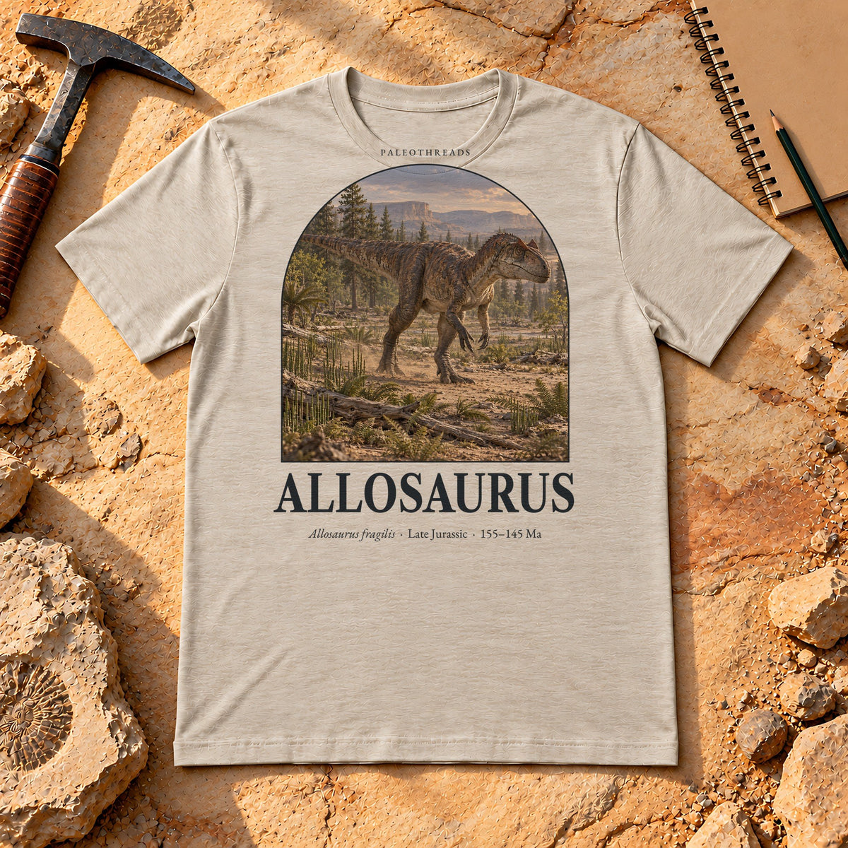 Allosaurus t-shirt flat lay – heather dust PaleoThreads paleo art tee on sandstone with a rock hammer
