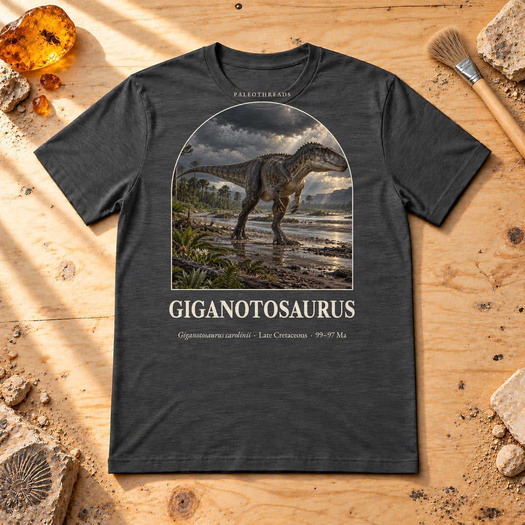 Giganotosaurus t-shirt flat lay – dark grey heather PaleoThreads paleo art tee on a fossil prep bench with amber