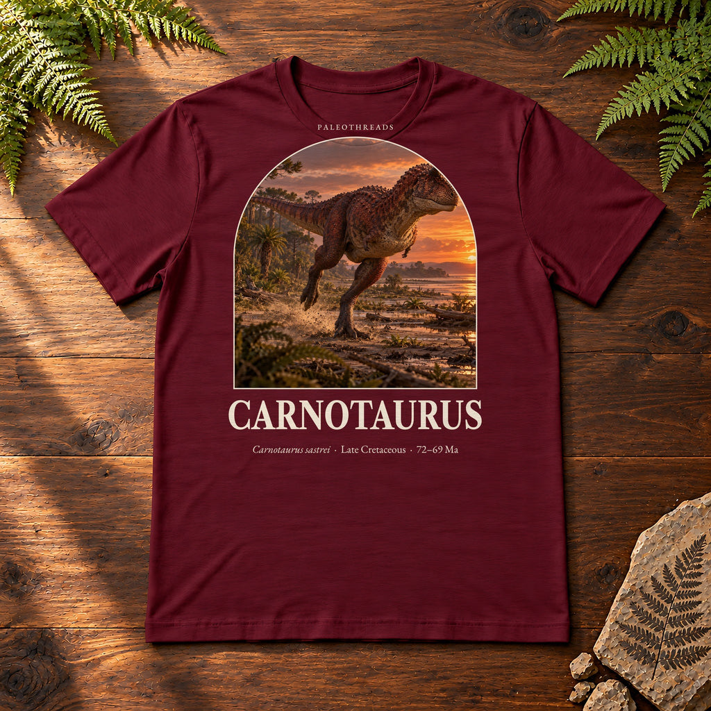 Carnotaurus t-shirt flat lay – maroon PaleoThreads paleo art tee on a walnut field desk with ferns