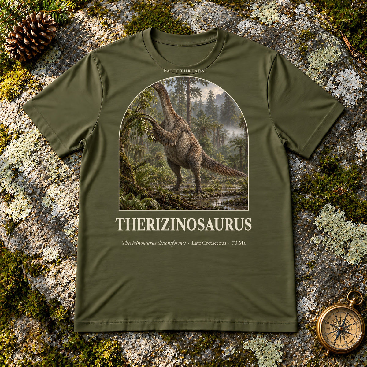 Therizinosaurus t-shirt flat lay – olive PaleoThreads paleo art tee on mossy rock with a brass compass