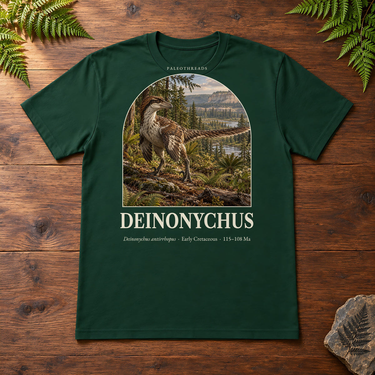 Deinonychus t-shirt flat lay – forest PaleoThreads paleo art tee on a walnut field desk with ferns