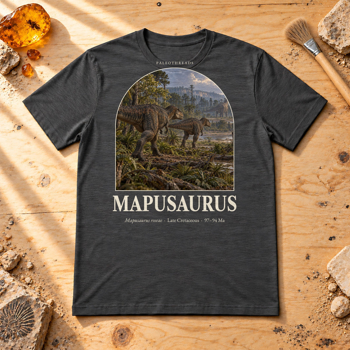 Mapusaurus t-shirt flat lay – dark grey heather PaleoThreads paleo art tee on a fossil prep bench with amber