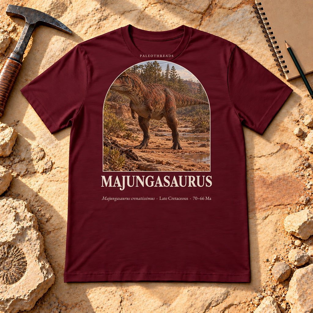 Majungasaurus t-shirt flat lay – maroon PaleoThreads paleo art tee on sandstone with a rock hammer