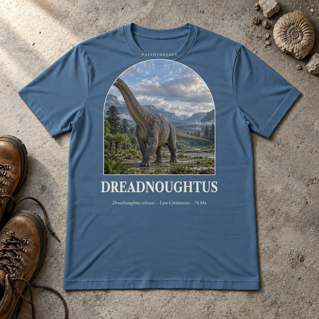 Dreadnoughtus t-shirt flat lay – steel blue PaleoThreads paleo art tee with hiking boots and an ammonite