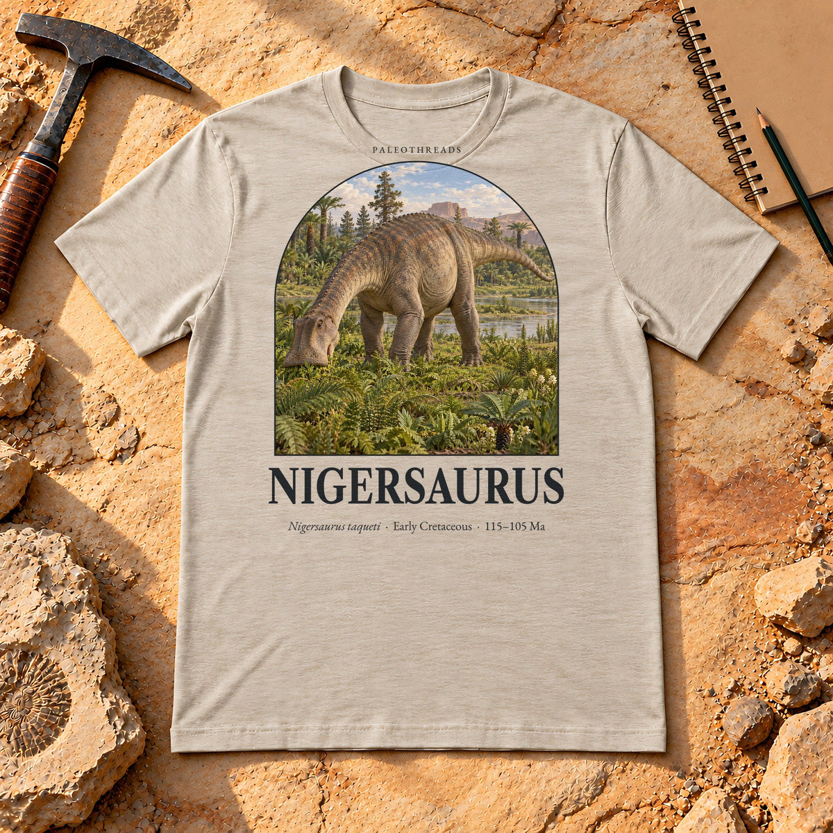 Nigersaurus t-shirt flat lay – heather dust PaleoThreads paleo art tee on sandstone with a rock hammer