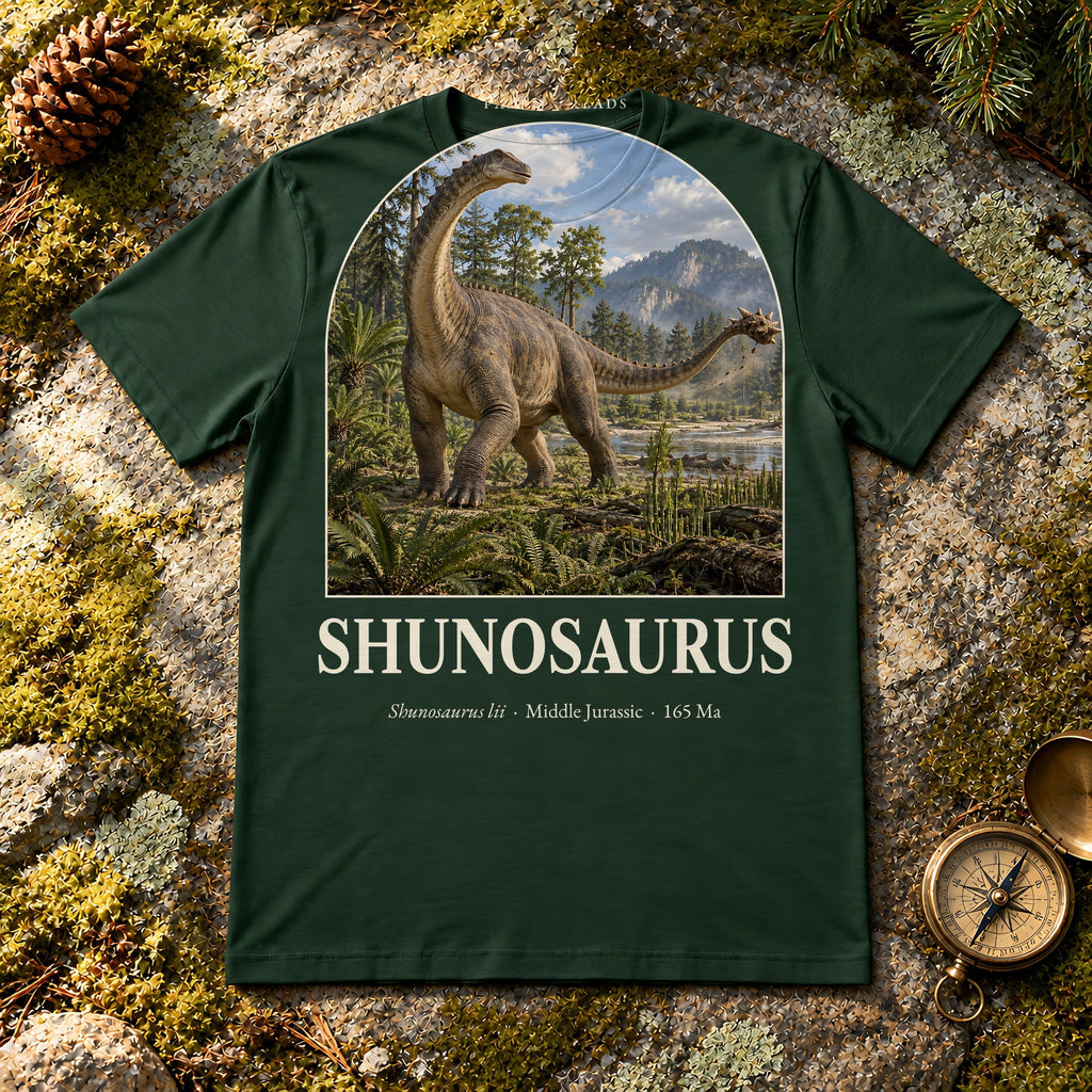 Shunosaurus t-shirt flat lay – forest PaleoThreads paleo art tee on mossy rock with a brass compass