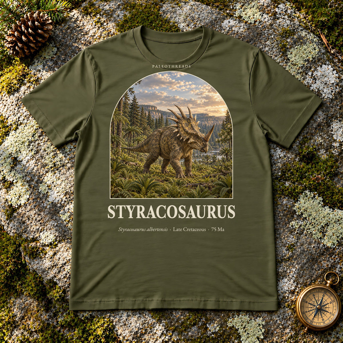 Styracosaurus t-shirt flat lay – olive PaleoThreads paleo art tee on mossy rock with a brass compass