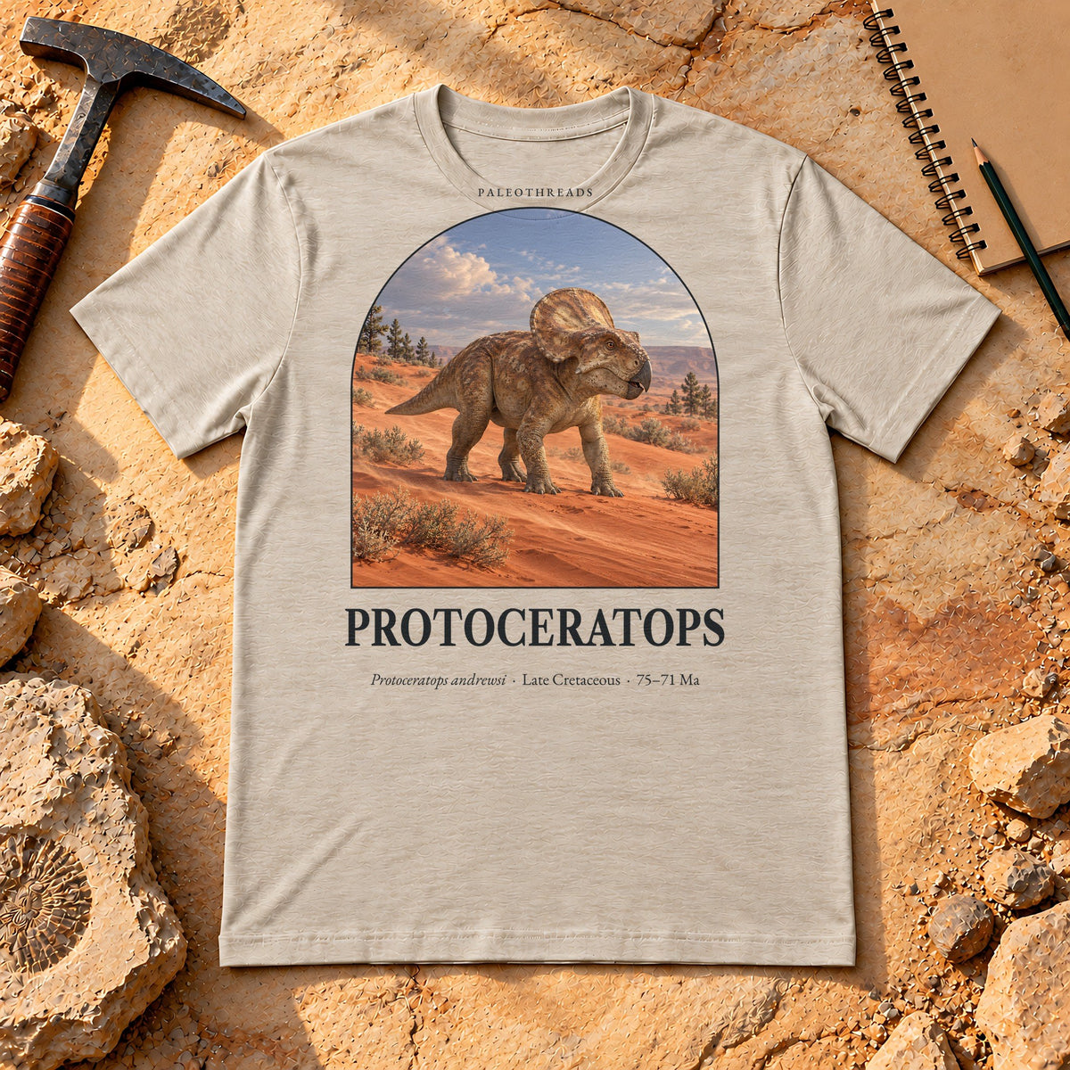 Protoceratops t-shirt flat lay – heather dust PaleoThreads paleo art tee on sandstone with a rock hammer