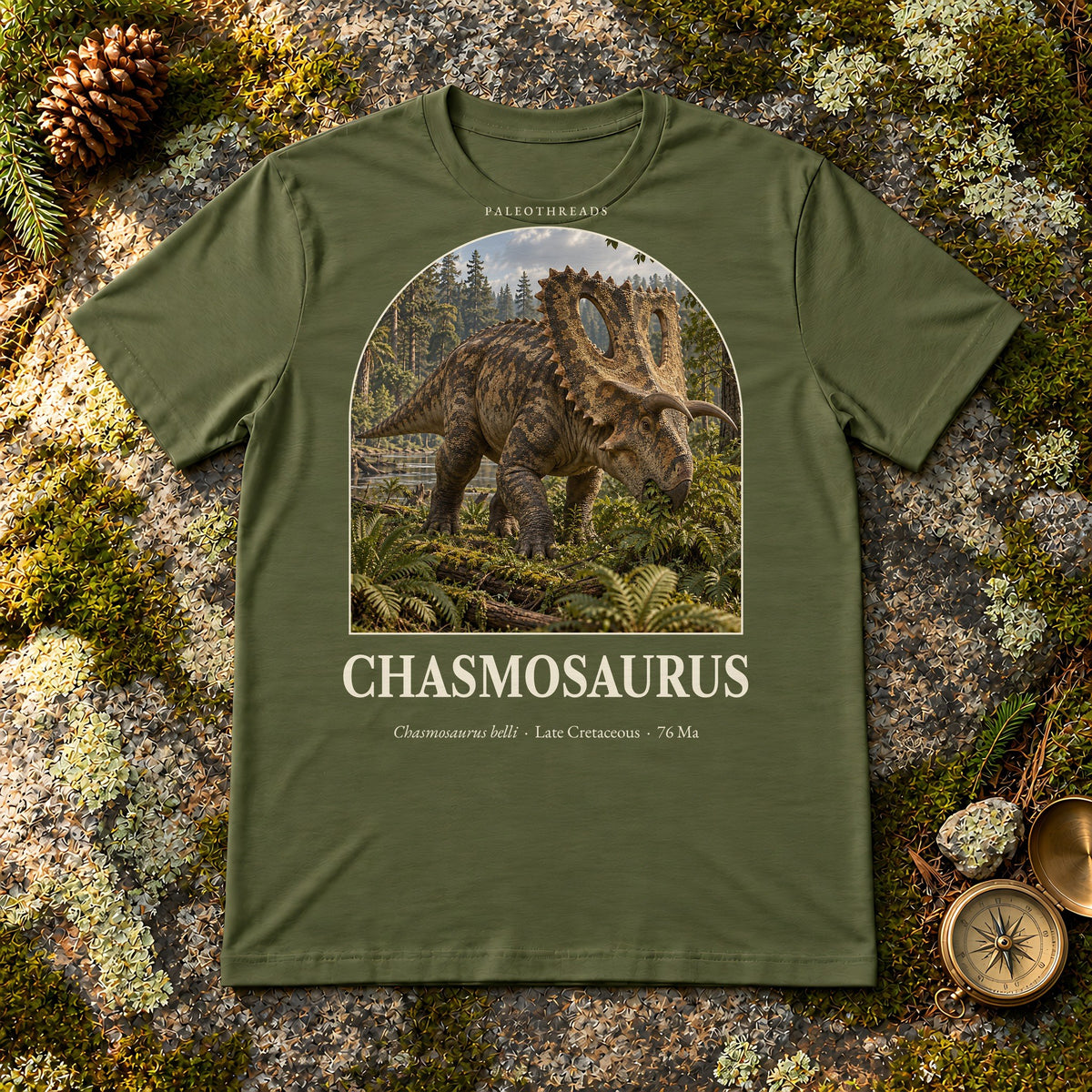 Chasmosaurus t-shirt flat lay – military green PaleoThreads paleo art tee on mossy rock with a brass compass
