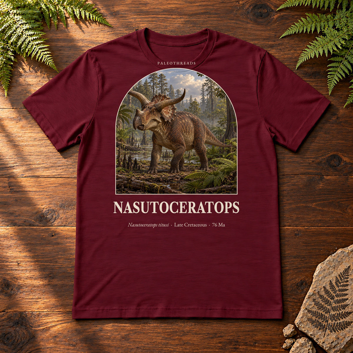 Nasutoceratops t-shirt flat lay – maroon PaleoThreads paleo art tee on a walnut field desk with ferns