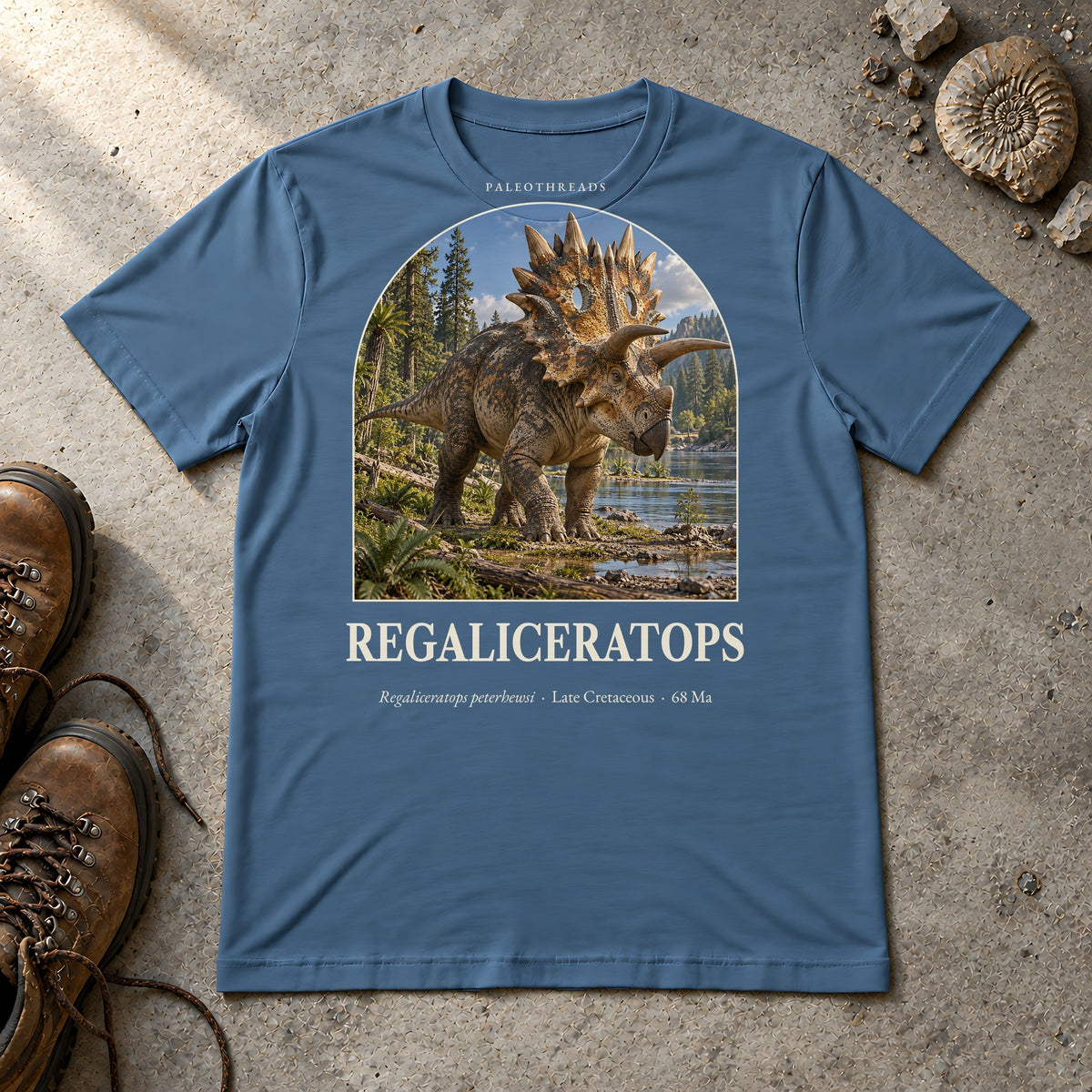Regaliceratops t-shirt flat lay – steel blue PaleoThreads paleo art tee with hiking boots and an ammonite