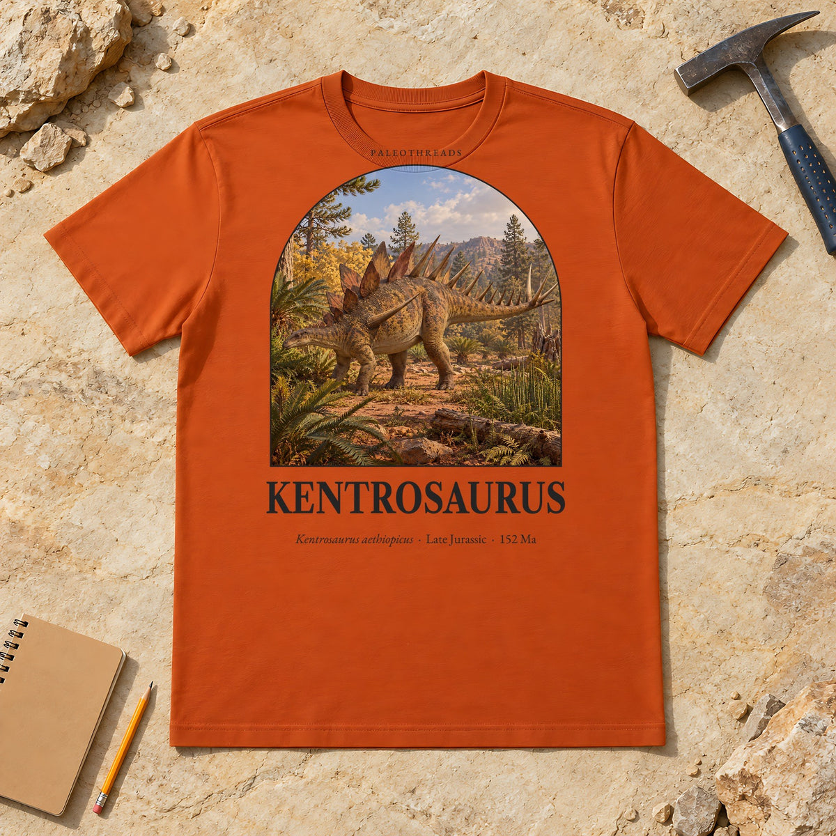Kentrosaurus t-shirt flat lay – autumn PaleoThreads paleo art tee on sandstone with a rock hammer