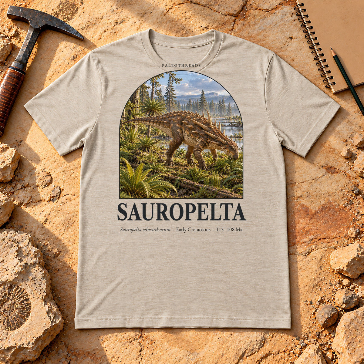 Sauropelta t-shirt flat lay – heather dust PaleoThreads paleo art tee on sandstone with a rock hammer