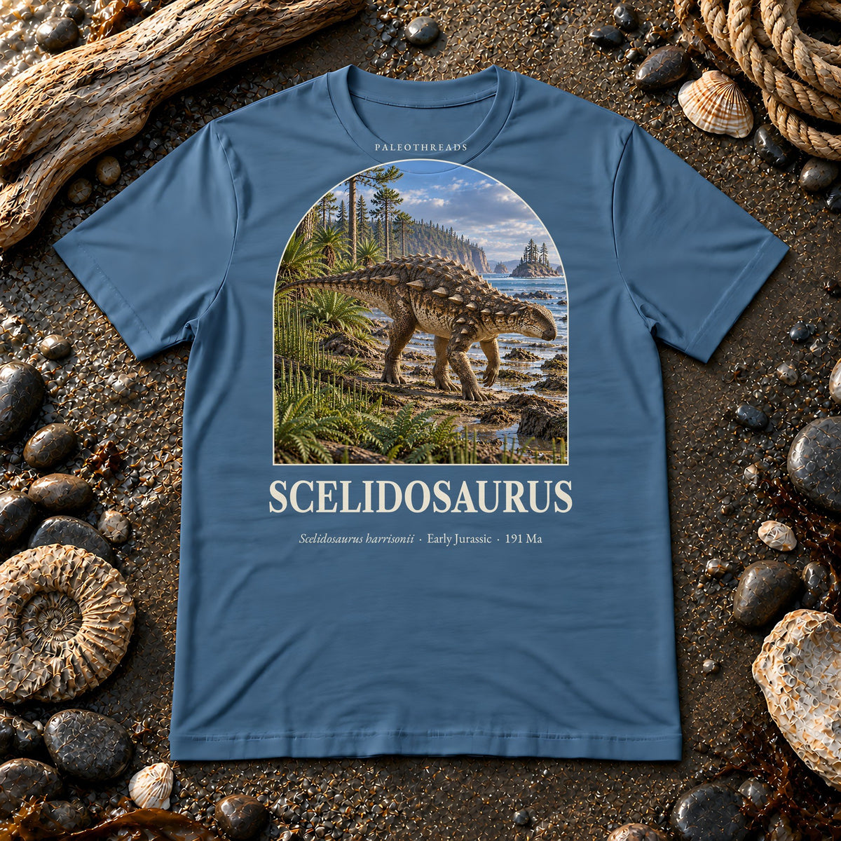 Scelidosaurus t-shirt flat lay – steel blue PaleoThreads paleo art tee on a fossil beach with driftwood