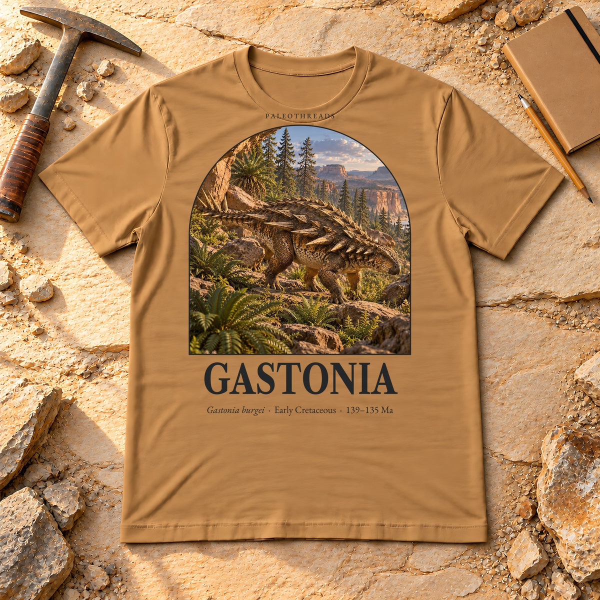 Gastonia t-shirt flat lay – toast PaleoThreads paleo art tee on sandstone with a rock hammer