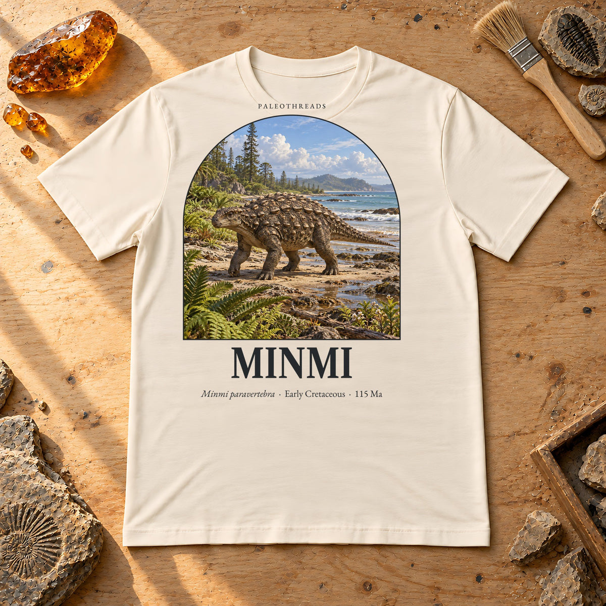 Minmi t-shirt flat lay – natural PaleoThreads paleo art tee on a fossil prep bench with amber