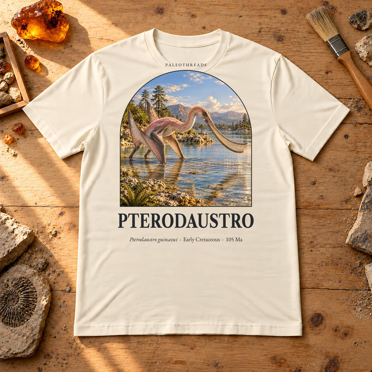 Pterodaustro t-shirt flat lay – soft cream PaleoThreads paleo art tee on a fossil prep bench with amber
