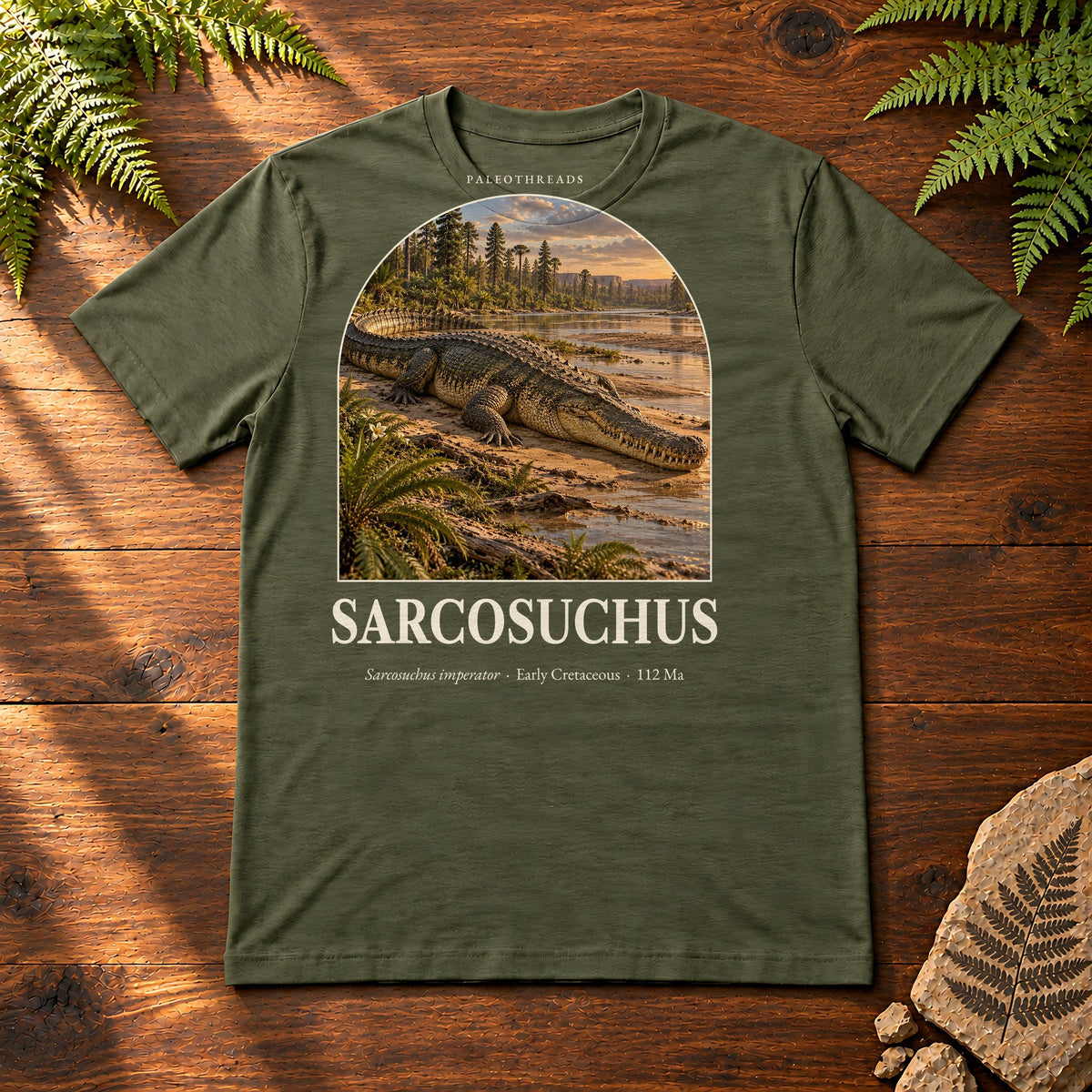 Sarcosuchus t-shirt flat lay – military green PaleoThreads paleo art tee on a walnut field desk with ferns