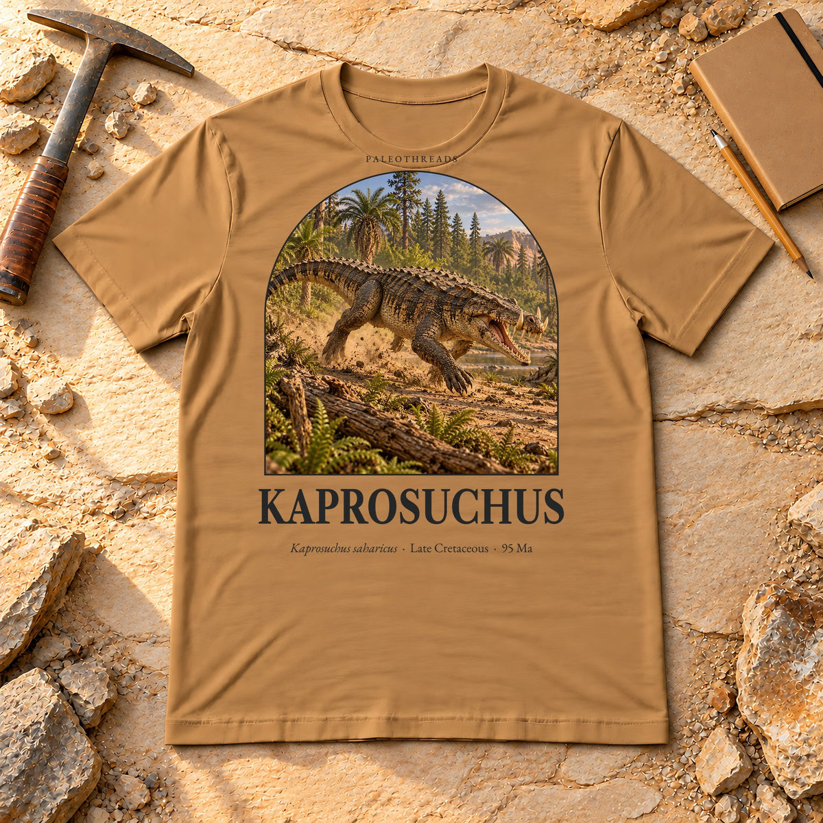 Kaprosuchus t-shirt flat lay – toast PaleoThreads paleo art tee on sandstone with a rock hammer