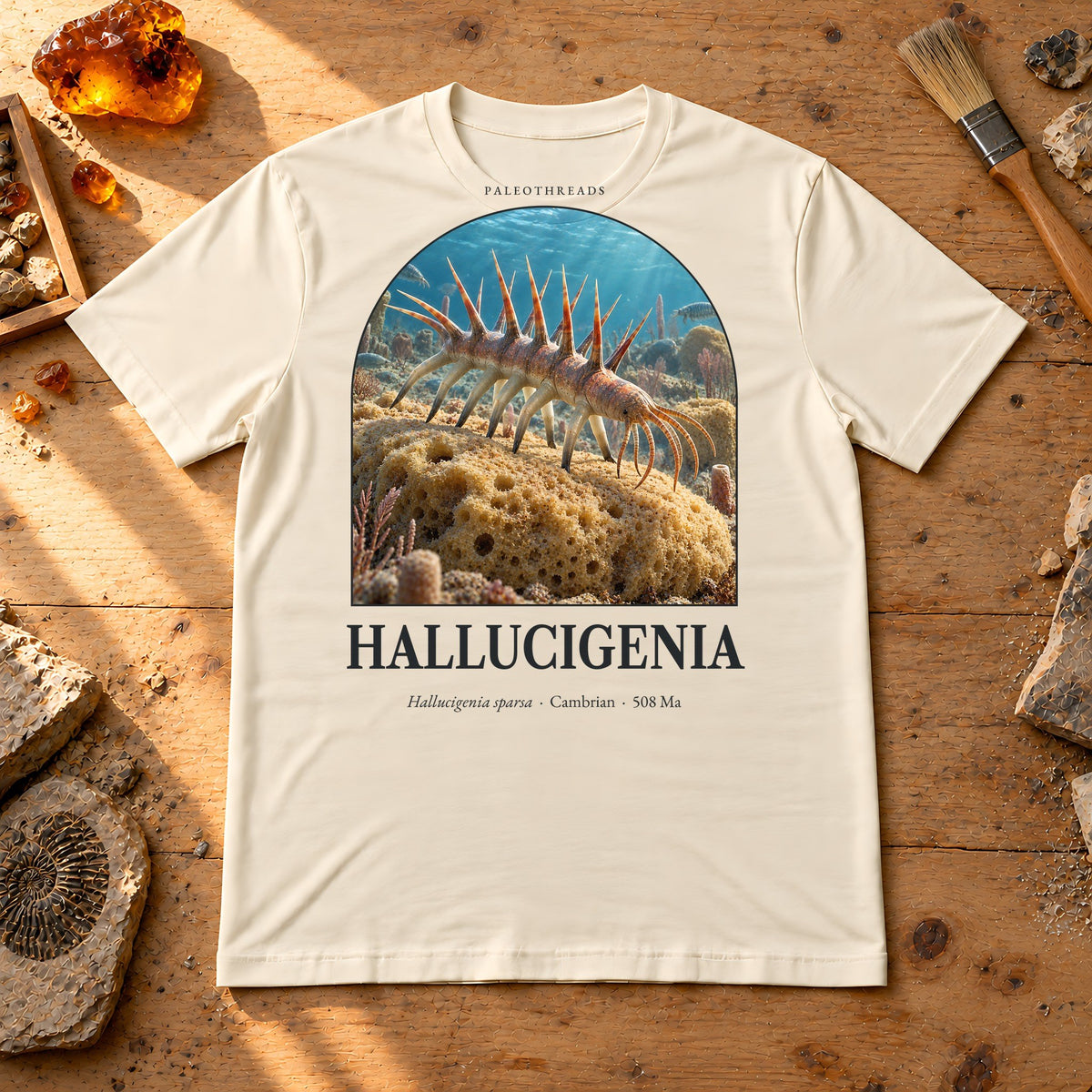 Hallucigenia t-shirt flat lay – soft cream PaleoThreads paleo art tee on a fossil prep bench with amber