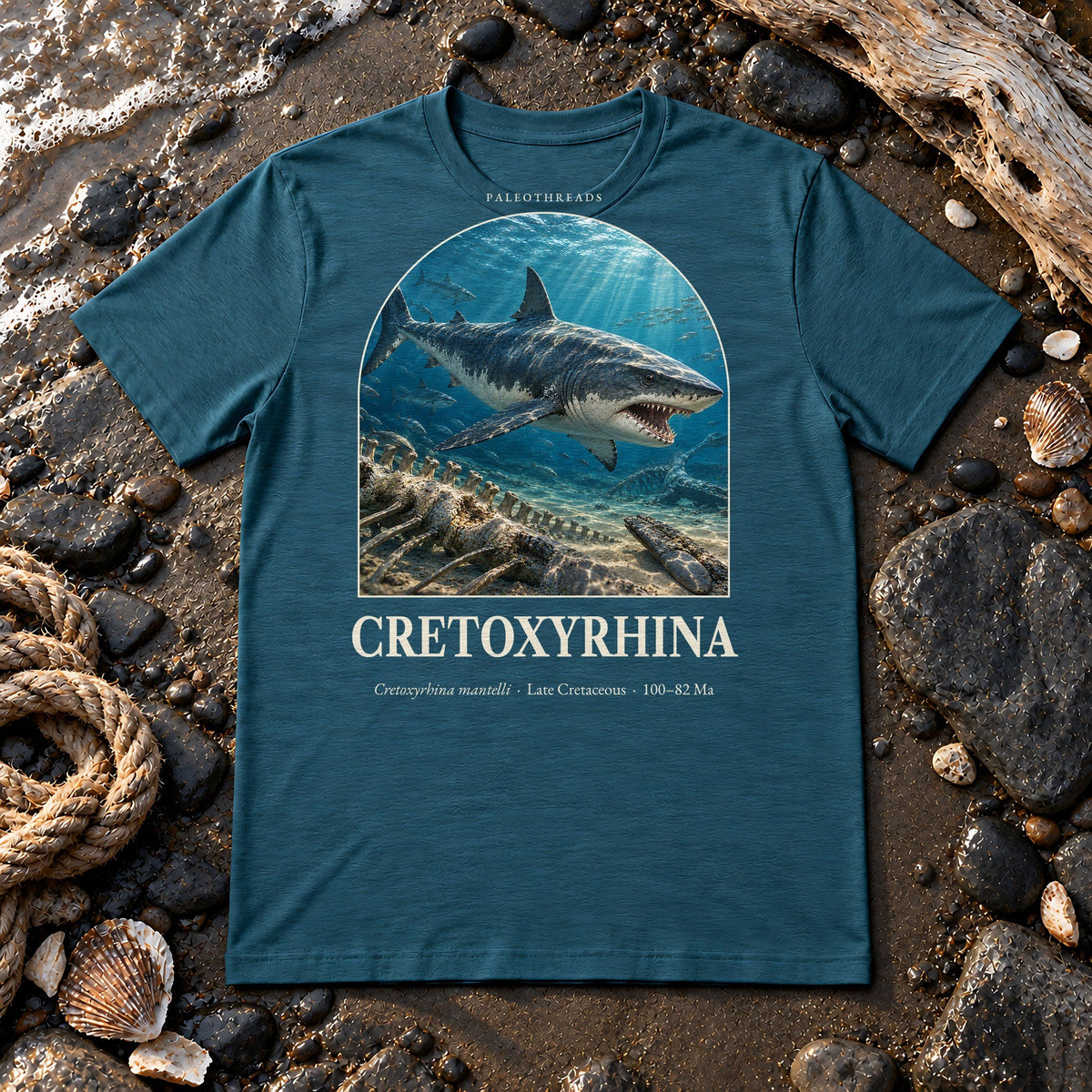 Cretoxyrhina t-shirt flat lay – heather deep teal PaleoThreads paleo art tee on a fossil beach with driftwood