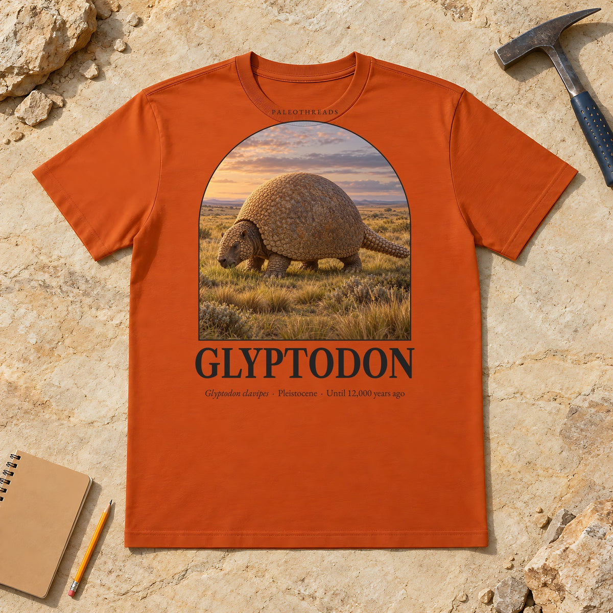 Glyptodon t-shirt flat lay – autumn PaleoThreads paleo art tee on sandstone with a rock hammer