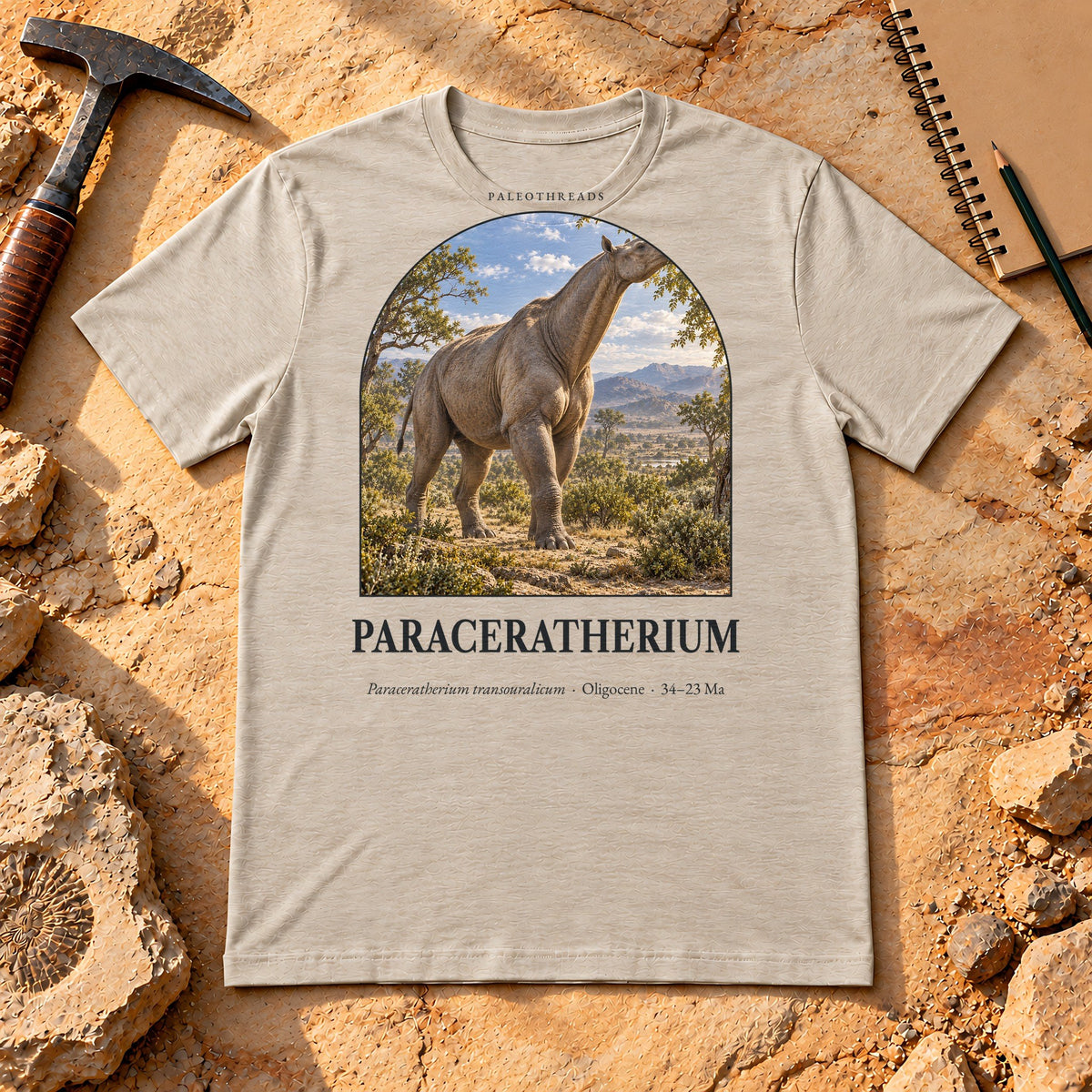 Paraceratherium t-shirt flat lay – heather dust PaleoThreads paleo art tee on sandstone with a rock hammer