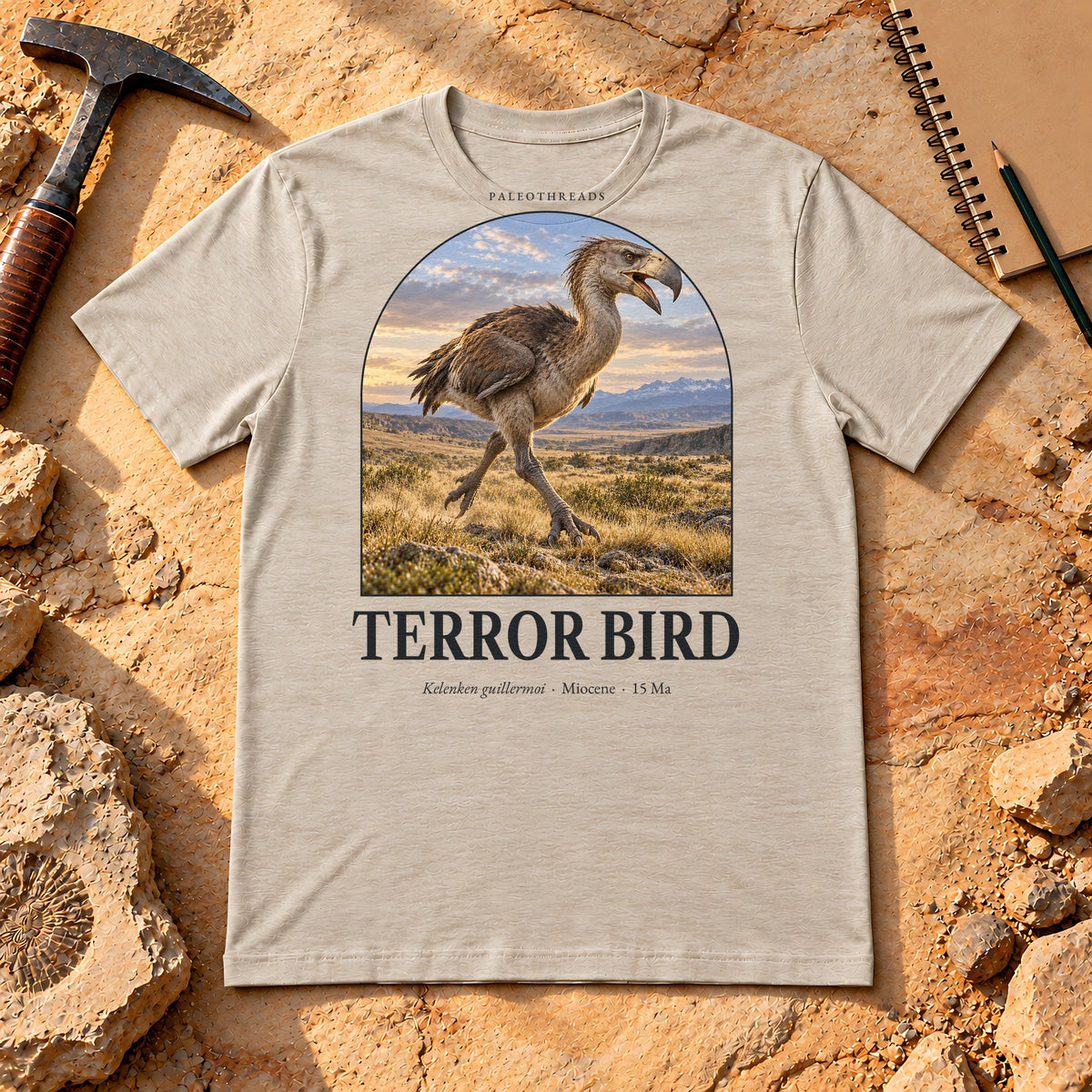 Terror Bird t-shirt flat lay – heather dust PaleoThreads paleo art tee on sandstone with a rock hammer